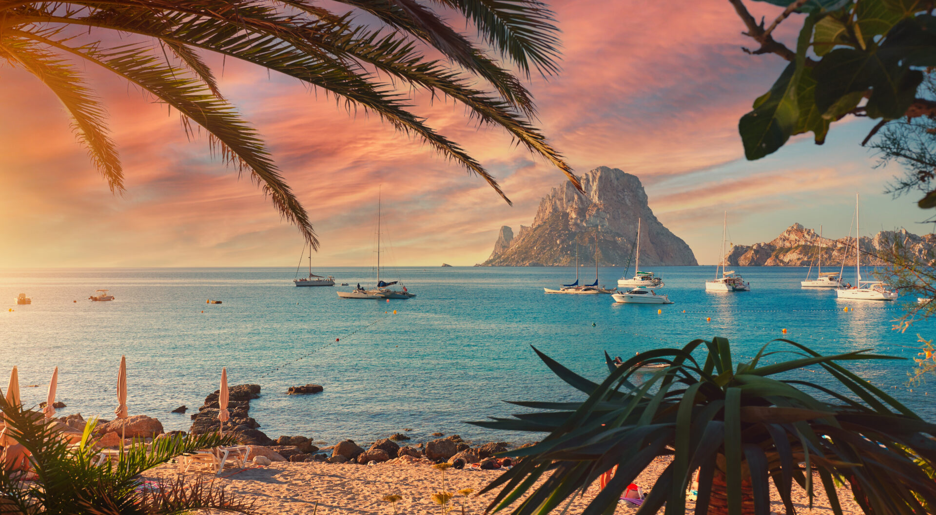 A large rock formation sitting out to sea with sail boats around, the photo is taken from the beach and the sun is setting wit pretty red and pink skies
