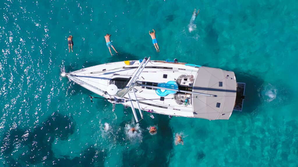 aerial view of a small white sailing boat, moored in crystal clear waters and showing people diving and jumping into the sea