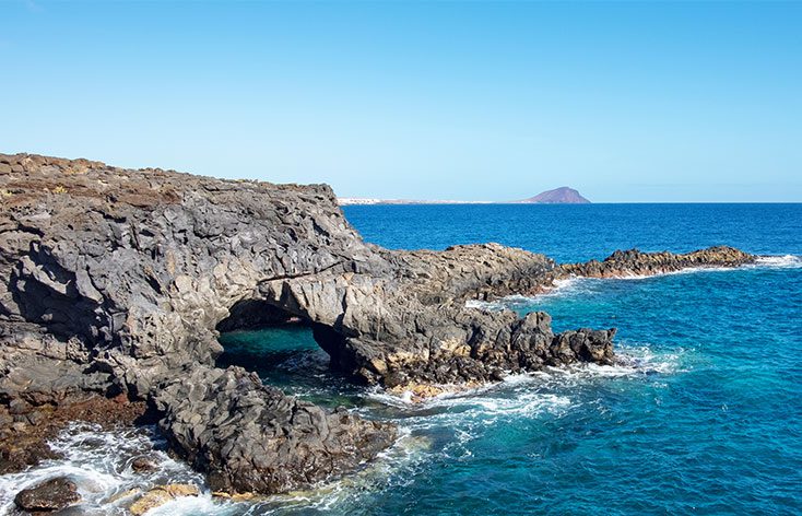 picture of black volcanic rocks at the edge of the sea, the naturel stones have formed into an archway and the water is all white where its crashing against the base of the rocks