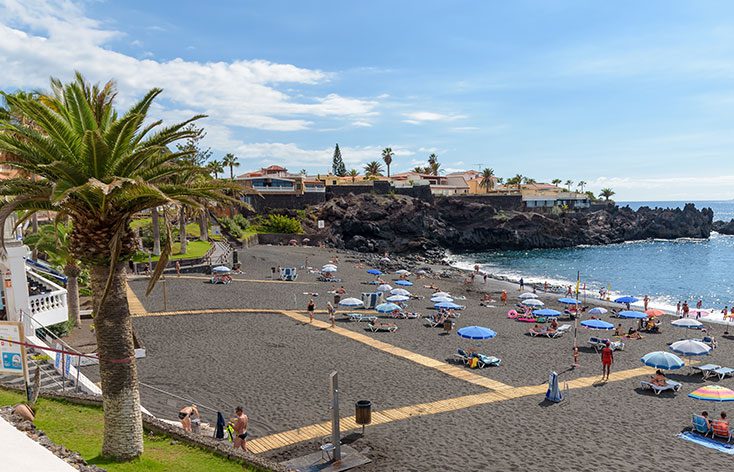 black volcanic sands at Puerto Santiago beach with sun bathers and dotted with parasols, palm trees and black volcanic rock formations