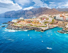 Aerial view of the resort of Puerto Santiago, blue seas crashing against the rocks, and small boats in the cove, mountains in the background